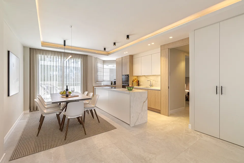 Fotografía de alquiler vacacional de un moderno comedor con mesa blanca, sillas elegantes y una cocina equipada al fondo en Nueva Andalucía