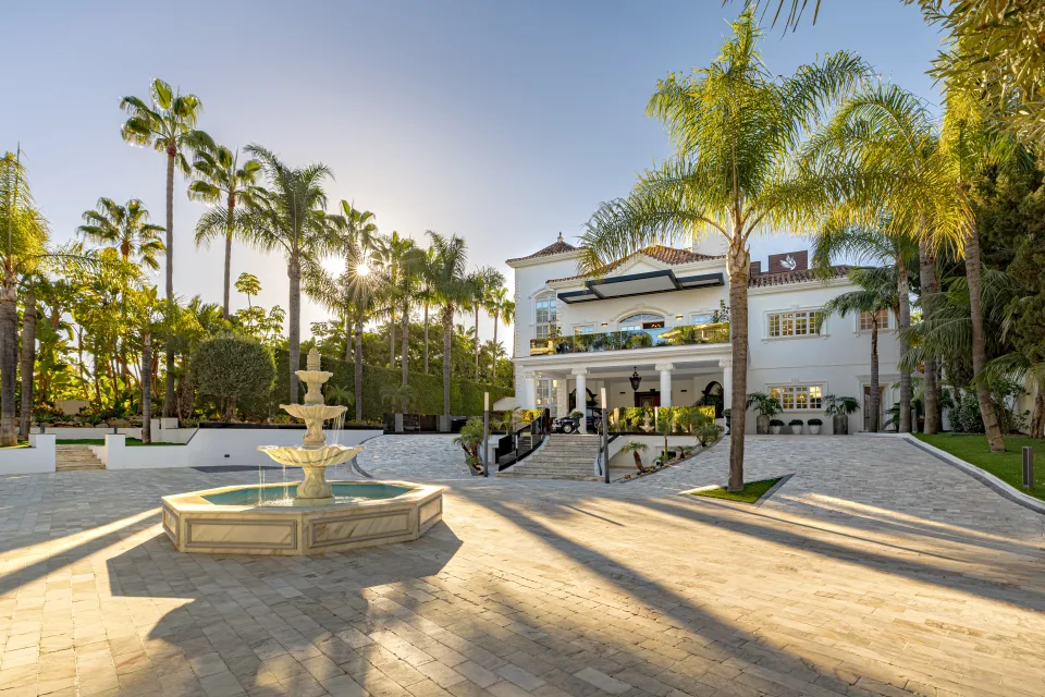 Fotografía de alquiler vacacional de un jardín con una fuente central rodeada de palmeras y una villa amplia al fondo en Marbella