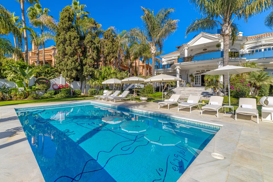 Fotografía de alquiler vacacional de una elegante villa con piscina rectangular, tumbonas y exuberante jardín rodeado de palmeras en Marbella