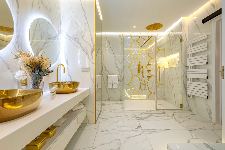 Fotografía de alquiler vacacional de un baño contemporáneo con mármol blanco, lavabos dorados y ducha de diseño en Marbella