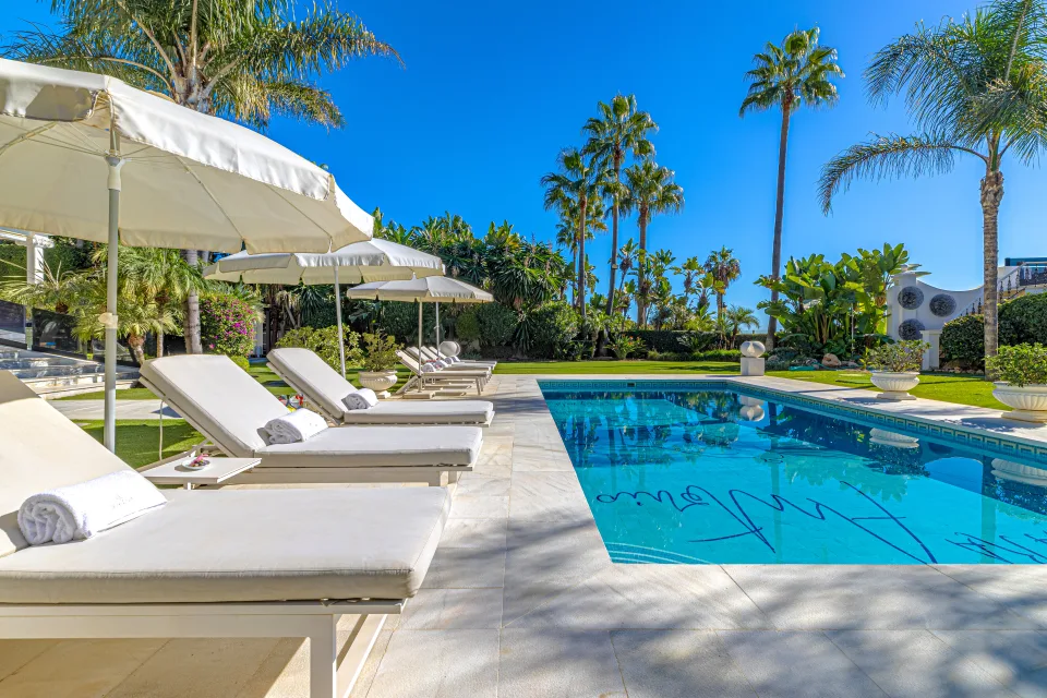 Fotografía de alquiler vacacional de una lujosa villa con piscina, tumbonas y palmeras en un entorno soleado en Marbella