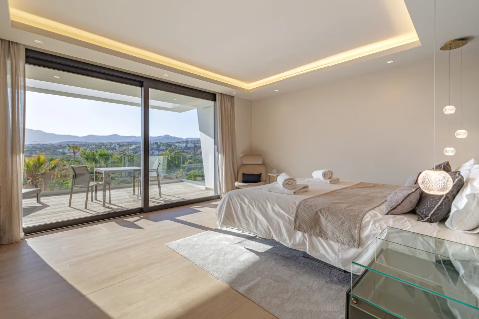 Fotografía de interiorismo y arquitectura de un dormitorio luminoso con cama amplia, acceso a terraza y vistas a la montaña en Estepona