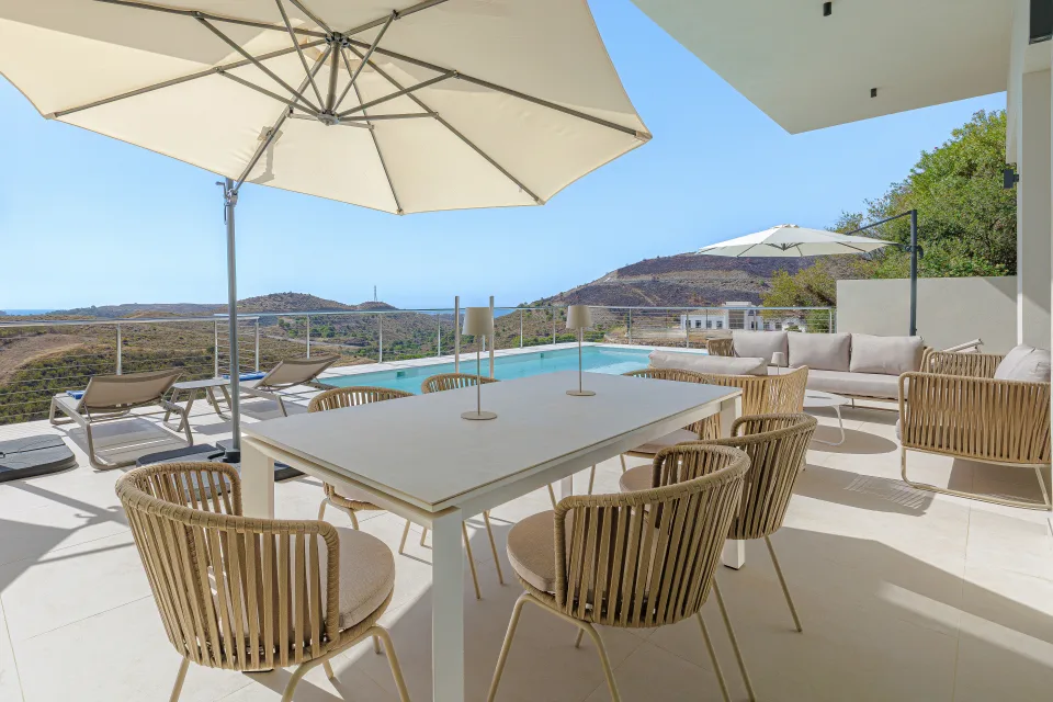 Fotografía de alquiler vacacional de una amplia terraza con mesas de comedor, sillas de mimbre y sombrillas junto a la piscina en Benahavís