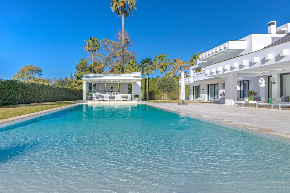 Fotografía de interiorismo y arquitectura de una villa moderna con piscina rodeada de palmeras y un área de descanso techada en Guadalmina Baja