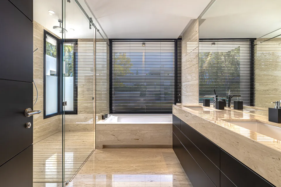 Fotografía de interiorismo y arquitectura de un baño moderno con acabados en mármol, mampara de vidrio y luz natural en Guadalmina Baja