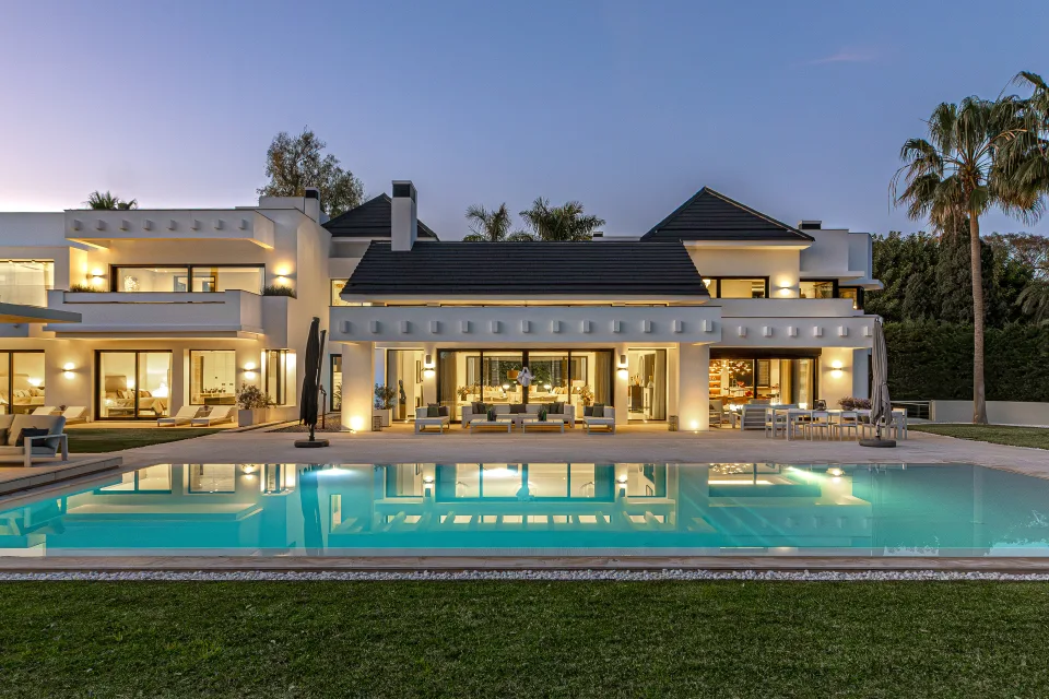 Fotografía de interiorismo y arquitectura de una villa moderna con piscina iluminada, amplios ventanales y jardín bien cuidado en Guadalmina Baja