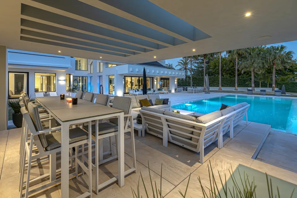Fotografía de interiorismo y arquitectura de un elegante porche con mobiliario de exterior, mesa grande y piscina iluminada en Guadalmina Baja
