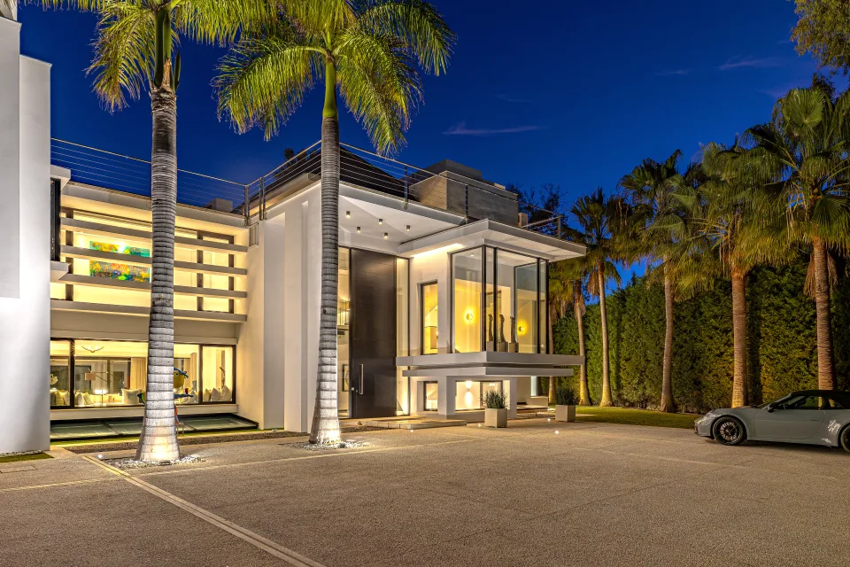 Fotografía de interiorismo y arquitectura de una villa elegante con fachada moderna, grandes ventanales y palmeras iluminadas en Guadalmina Baja