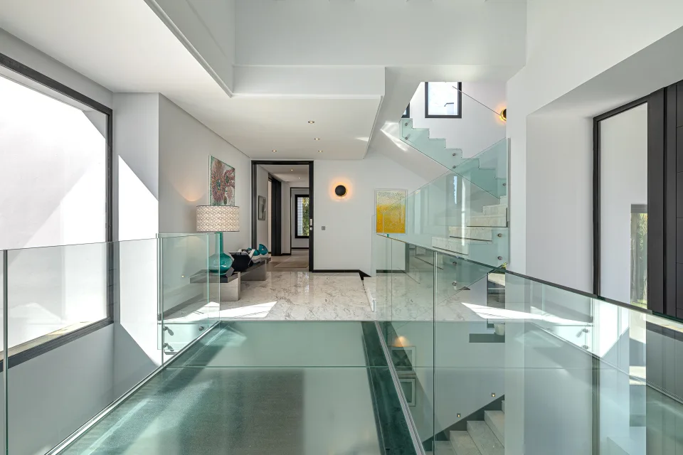 Fotografía de interiorismo y arquitectura de un amplio vestíbulo con escaleras de vidrio, luces modernas y mobiliario elegante en Guadalmina Baja