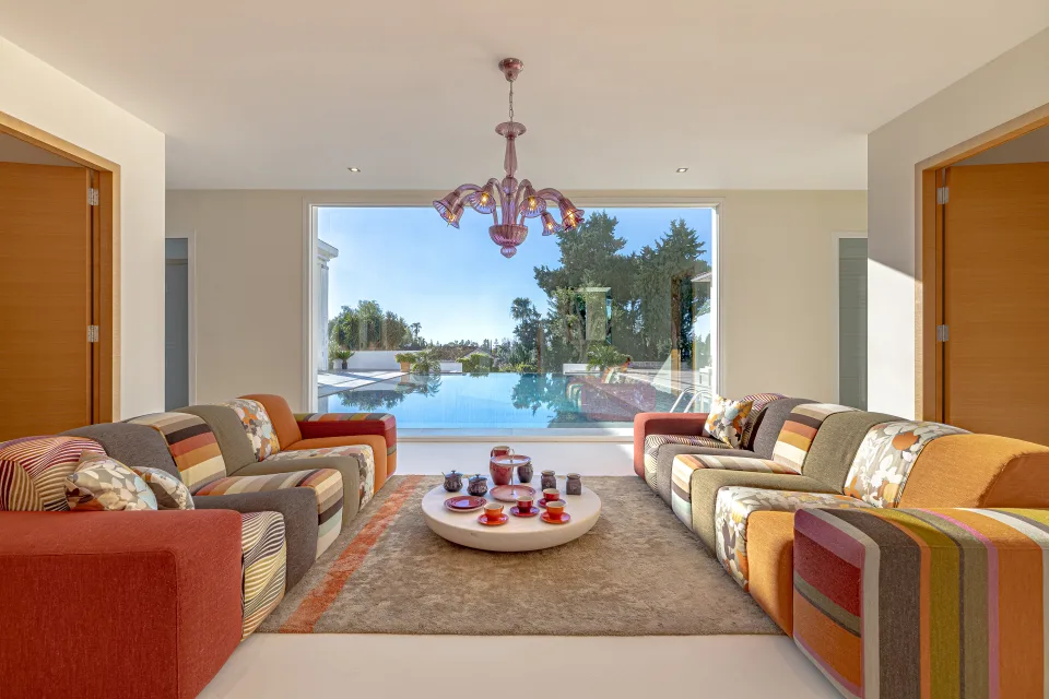 Real estate photograph of a bright lounge with multicoloured sofas, round coffee table and views of the pool in Marbella