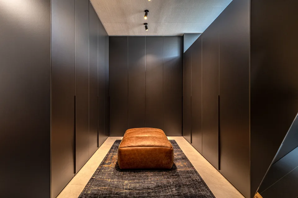 Fotografía inmobiliaria de un vestidor moderno con armarios empotrados oscuros y un ottoman de cuero marrón en Marbella