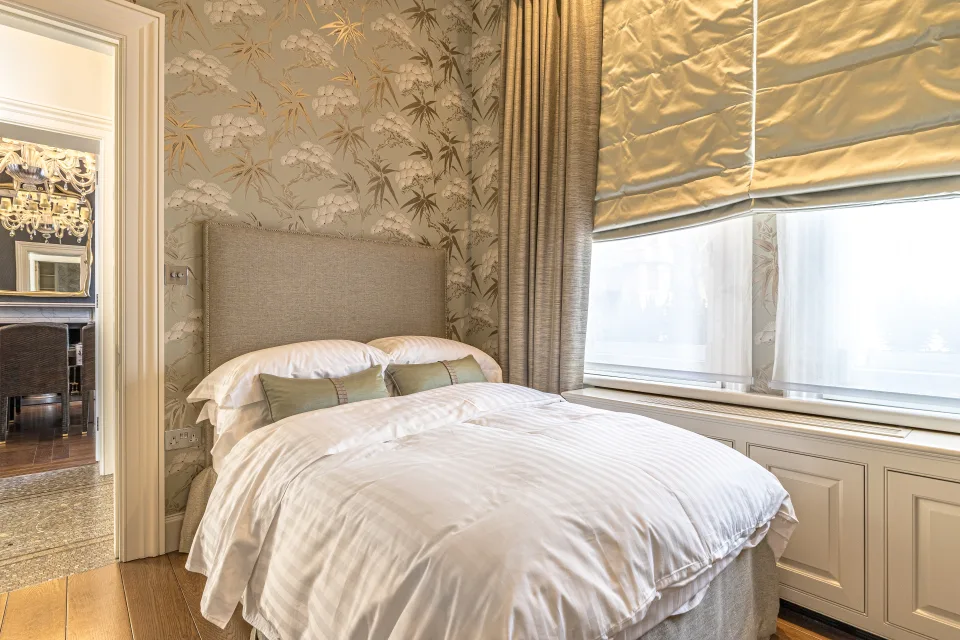 Fotografía inmobiliaria de un dormitorio luminoso con papel pintado floral, cama doble con sábanas blancas y cortinas elegantes en Londres